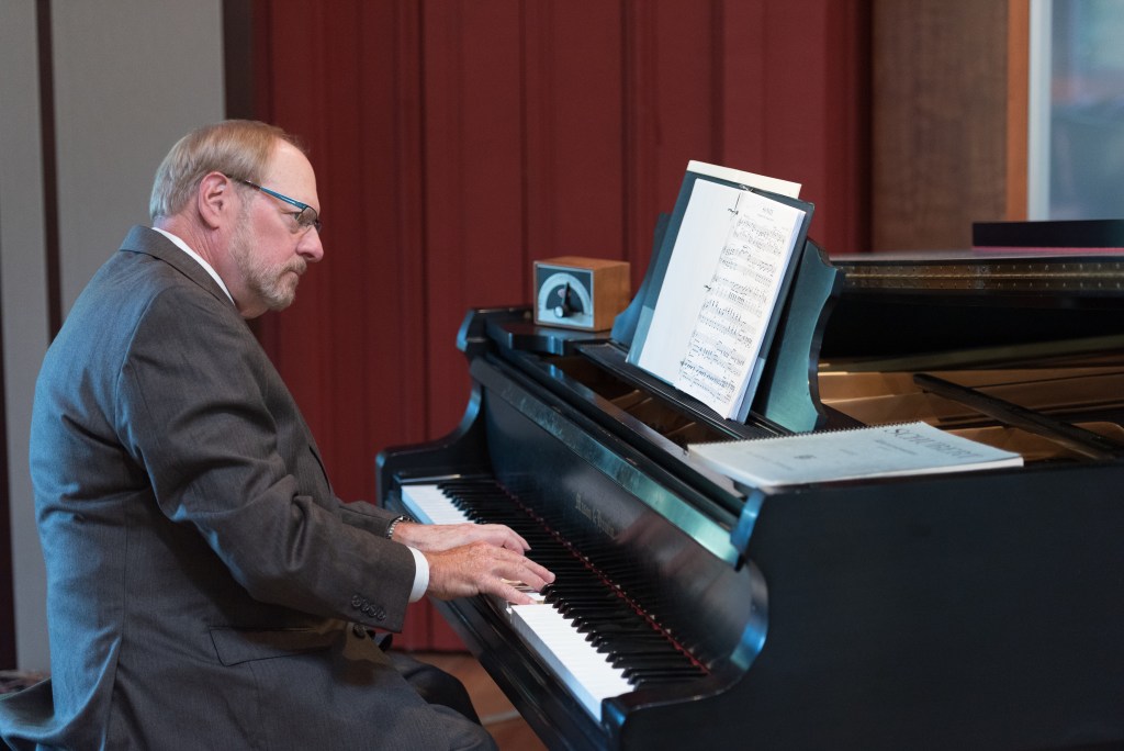 Medium long shot. George Gopen, a white man with short reddish greying hair, mustache, and beard. He is wearing glasses in a dark grey jacket and trousers with a white collared shirt. He is sitting at a black grand piano—his hands on the keys. There are a metronome and sheets of music on top of the piano. There are deep red drapes and the edge of a large window visible in the background.
