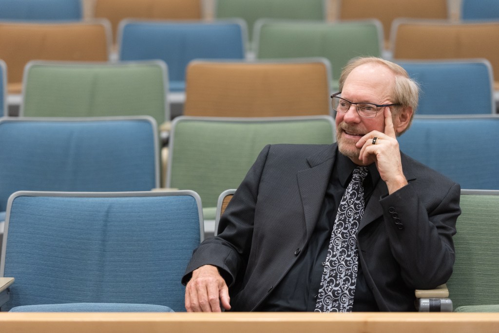 Medium wide shot of George Gopen, professional writing consultant — a smiling, older white man in an auditorium. He is wearing a black suit with an ornate tie with white designs. He has short, reddish grey hair, mustache, and beard. He has glasses. His head is resting in his left hand and he is gazing to the left. The seats around him are empty and alternate in colors from blue to green to orange.