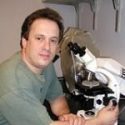 Medium shot of Adam, a white man with short brown hair in a green Tshirt sitting at a powerful looking white microscope, his hand on the focus knob.