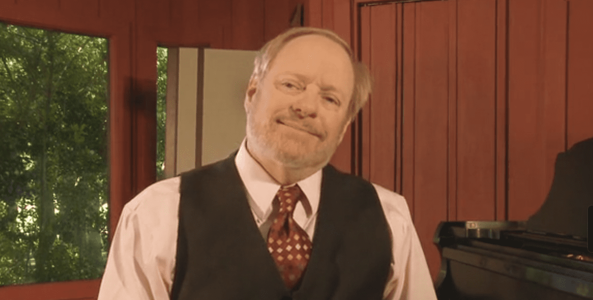 George Gopen, a smiling white man with short reddish grey hair, beard, and mustache sitting at a grand piano.