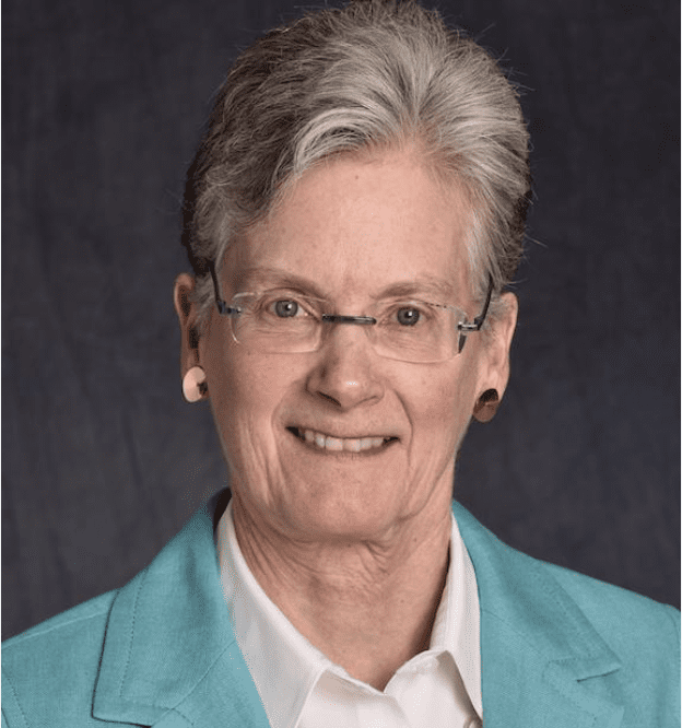 White woman, short grey hair, silver glasses, turquoise blazer, white blouse, smiling,